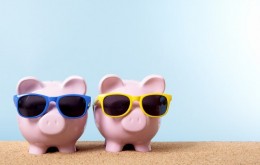 Two pink piggy banks on a beach with sunglasses.  Space for copy.  Studio shot with plain blue background - warm color and directional lighting are intentional.